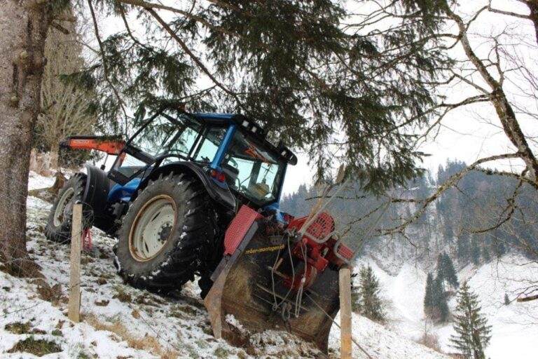 Ein Traktor steht in einem schneebedeckten Hang, bereit, eine Pflügmaschine bei der Verlegung von Leitungen zu unterstützen.