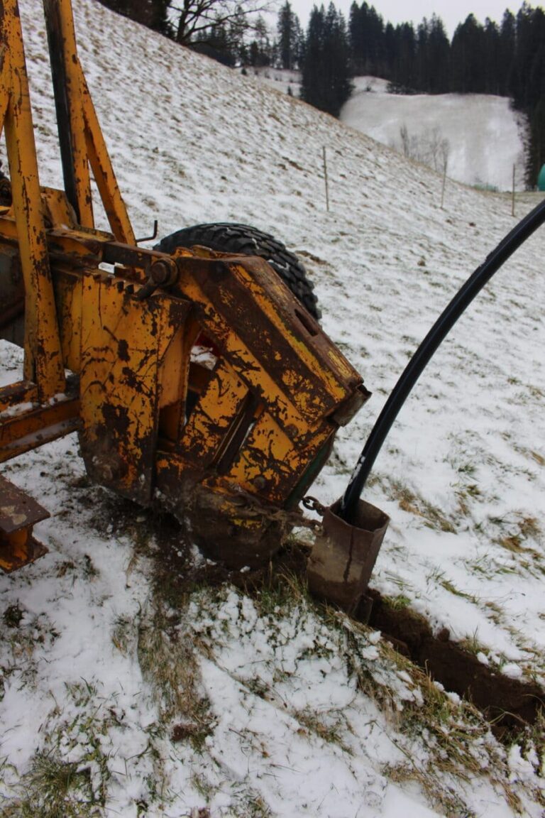 Eine Pflügmaschine arbeitet auf dem mit Schnee bedeckten Feld, während sie eine Wasserleitung im Boden verlegt. Die Pflugschare durchbrechen die winterliche Erde, um den Weg für die neue Leitung zu bahnen.